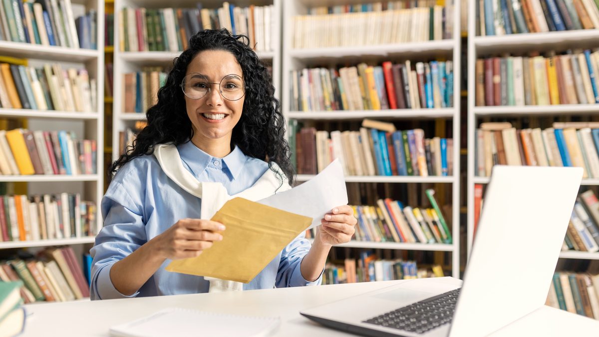 Une jeune femme souriante aux cheveux bouclés, devant un ordinateur portable et des étagères de livres, tient une enveloppe et des documents