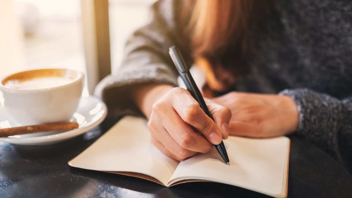 Femme écrivant dans un carnet posé sur une table, à côté d'une tasse de café